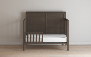 A Child Craft Toddler Guard Rail in dapper gray, crafted from sturdy wood with a paneled headboard, partial guard rail, and white mattress, sits against a light wall on a light wood floor.