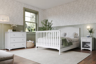 A bright, cozy bedroom with a white full-size bed featuring Child Craft F06401 Full-Size Bed Rails in matte white, a matching nightstand and dresser, green and white walls with leafy wallpaper, lots of plants, a large window, and soft natural light.