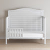 The Child Craft F09501 Toddler Guard Rail in matte white features a sturdy wood build, slatted back and sides, and a simple design. It sits on a light wood floor against a plain beige wall, adding clean style to any toddler's room.