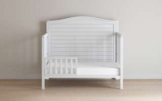 The Child Craft F09501 Toddler Guard Rail in matte white features a sturdy wood build, slatted back and sides, and a simple design. It sits on a light wood floor against a plain beige wall, adding clean style to any toddler's room.