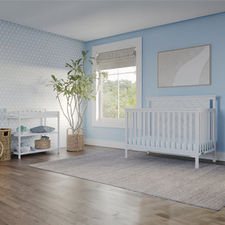 A bright nursery with light blue walls, a Child Craft Hampton Flat Top 4-in-1 Convertible Crib in matte white, a changing table with storage baskets, a potted plant, a large window with a roman shade, and a neutral area rug on wooden floors.