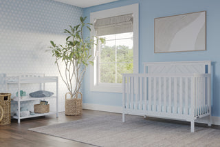 A bright, airy nursery with light blue walls features the Child Craft Hampton Flat Top 4-in-1 Convertible Crib in matte white, a matching changing table, large window, potted plant, and soft gray rug on dark wood floors for a calm, inviting feel.