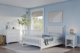 Bright, airy bedroom with light blue walls, white bed, nightstand, and dresser. The Child Craft Hampton Flat Top 4-in-1 Convertible Crib in matte white sits by the window near a large plant; sunlight highlights the cozy rug and minimalist decor.