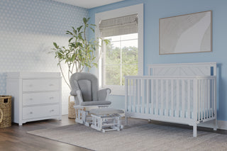 A nursery with light blue walls, a Child Craft Hampton Flat Top 4-in-1 Convertible Crib in Matte White, a gray cushioned rocking chair and ottoman, a white dresser, large plant, woven baskets, a window, and framed abstract artwork.
