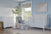 A nursery with light blue walls, a Child Craft Hampton Flat Top 4-in-1 Convertible Crib in Matte White, a gray cushioned rocking chair and ottoman, a white dresser, large plant, woven baskets, a window, and framed abstract artwork.