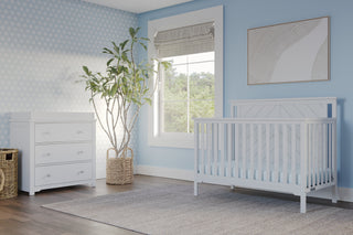A bright nursery with light blue walls, the Child Craft Hampton Flat Top 4-in-1 Convertible Crib in Matte White, a white dresser with baskets, a large potted plant, a window with a roman shade, and framed artwork above the crib.