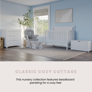 A nursery with light blue walls, white furniture including the Child Craft Hampton Flat Top 4-in-1 Convertible Crib in Matte White, a dresser, chest, gray glider chair with ottoman, window blinds, and a beige rug on wooden floors. Classic Cozy Cottage.