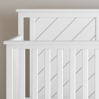 A close-up of the Child Craft Hampton Flat Top 4-in-1 Convertible Crib in matte white, crafted from sturdy pine wood with diagonal slats on the headboard and side railings, shown against a beige wall.