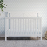 The Child Craft Hampton Flat Top 4-in-1 Convertible Crib in Matte White, crafted from sturdy pine with non-toxic finishes, stands by a floral blue wall, accompanied by a potted plant and woven basket on a wooden floor.