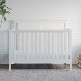 The Child Craft Hampton Flat Top 4-in-1 Convertible Crib in Matte White, crafted from sturdy pine with non-toxic finishes, stands by a floral blue wall, accompanied by a potted plant and woven basket on a wooden floor.