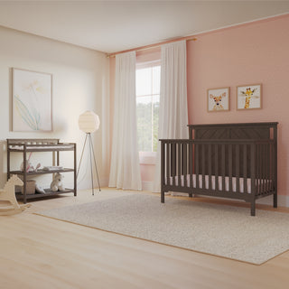 A nursery featuring the Child Craft Hampton Flat Top 4-in-1 Convertible Crib in Dapper Gray, a changing table with shelves, a rocking horse, animal art on a pink polka-dot accent wall, light curtains by the window, and a soft rug on the floor.