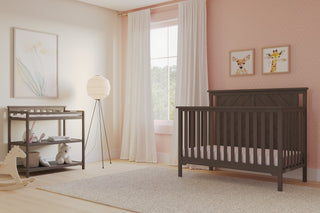 A cozy nursery with a dark wooden crib, Child Craft Flat Top Baby Changing Table in Dapper Gray with a baby changing pad, rocking horse, soft rug, floor lamp, pastel pink polka dot walls, animal art above the crib, and sunlight through white curtains.