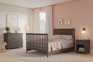 A cozy bedroom featuring the Child Craft Hampton Flat Top 4-in-1 Convertible Crib in Dapper Gray, with gray bedding, matching dresser and nightstand, a lamp, wall art, white-curtained window, beige rug, and pink polka dot wallpaper.