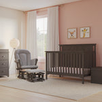 A nursery with light pink polka dot wallpaper, a Child Craft Hampton Flat Top 4-in-1 Convertible Crib in Dapper Gray, matching dresser and toy chest, a gray glider chair with ottoman, rocking horse, animal art on the walls, and sunlight streaming in.