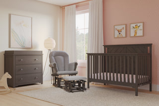 A cozy nursery with pink walls features the Child Craft Hampton Flat Top 4-in-1 Convertible Crib in Dapper Gray, a matching dresser, gray glider chair with ottoman, rocking horse, animal prints, pastel artwork, and white curtains by a large window.