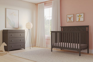 A modern nursery with a Child Craft Hampton Flat Top 4-in-1 Convertible Crib in Dapper Gray, a matching dresser, floor lamp, window with white curtains, pink polka dot walls with animal prints, and a small rocking horse on light wood floors.