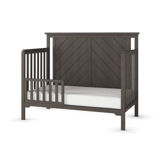 The Child Craft F09501 Toddler Guard Rail in Dapper Gray features sturdy wood construction, open slatted sides, and a chevron-patterned headboard, displayed with a white mattress against a white background.