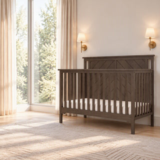 Child Craft Hampton Flat Top 4-in-1 Convertible Crib in Dapper Gray, featuring a flat top and chevron detail on the headboard, placed in a room next to a window.