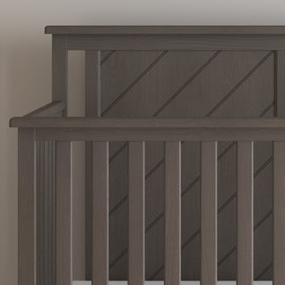 A close-up of the Child Craft Hampton Flat Top 4-in-1 Convertible Crib in Dapper Gray, featuring vertical slats and a diagonal slat pattern on the headboard, set against a light-colored wall.