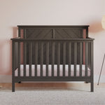 The Child Craft Hampton Flat Top 4-in-1 Convertible Crib in Dapper Gray features vertical slats and a geometric headboard design, set on a light wooden floor against a pink polka dot wall, with a lamp and a gray rug nearby.