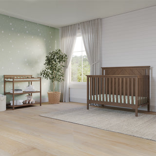 A modern nursery with the Child Craft Hampton Flat Top 4-in-1 Convertible Crib in Dusty Heather, a matching changing table, green accent wall with white stars, large window with sheer curtains, potted plant, and beige rug on light wood flooring.