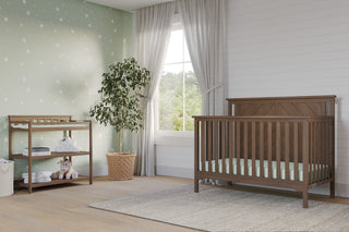 A nursery with a wooden crib, Child Craft Flat Top Baby Changing Table in Dusty Heather, storage baskets, a plush unicorn toy on the shelf, potted plant, light wood floor, gray rug, green wall with white stars, and gray curtains by the window.