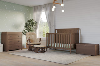 A nursery room with the Child Craft Hampton Flat Top 4-in-1 Convertible Crib in Dusty Heather, adjustable mattress support, a dresser, storage chest, beige rocking chair with ottoman, potted plant, rug, wood flooring, and star wallpaper.