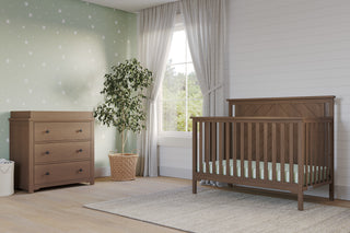 A modern nursery featuring the Child Craft Hampton Flat Top 4-in-1 Convertible Crib in Dusty Heather with a matching dresser, potted plant, light green star wall, white curtains, large window, and a light gray rug on wood floors.