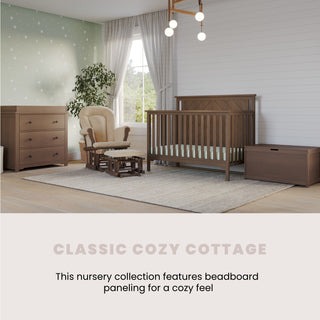 A cozy nursery with sturdy pine furniture, including the Child Craft Hampton Flat Top 4-in-1 Convertible Crib in Dusty Heather, dresser, chest, and rocking chair on a rug. Sunlight pours in, highlighting green dotted wallpaper and beadboard. Classic Cozy Cottage.