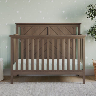 The Child Craft Hampton Flat Top 4-in-1 Convertible Crib in Dusty Heather with a chevron headboard sits on a light rug. A pale green wall with stars, potted plant, and wicker basket create a calm nursery ambiance.