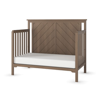 The Child Craft Hampton Flat Top 4-in-1 Convertible Crib in Dusty Heather features vertical side slats, an open side, a chevron-patterned headboard, and a white mattress, all crafted in a classic pine wood style.