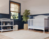A bright nursery showcases the Child Craft Hampton Arch Top 4-in-1 Convertible Crib in matte white with adjustable mattress support, a changing table with toys and books, potted plant, basket, floor lamp, window with wooden blinds, blue wainscoting, and patterned wallpaper.