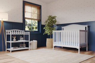 A bright nursery showcases the Child Craft Hampton Arch Top 4-in-1 Convertible Crib in matte white with adjustable mattress support, a changing table with toys and books, potted plant, basket, floor lamp, window with wooden blinds, blue wainscoting, and patterned wallpaper.