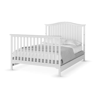 The Child Craft Hampton Arch Top 4-in-1 Convertible Crib in Matte White features vertical slats on the headboard and footboard, adjustable mattress support, and is shown with white bedding and a gray blanket.