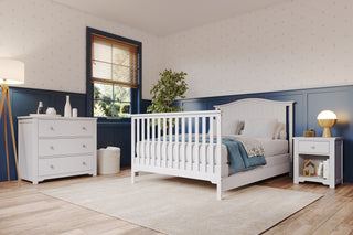 A cozy bedroom showcases the Child Craft Hampton Arch Top 4-in-1 Convertible Crib in matte white, paired with a matching nightstand and dresser. Blue wainscoting, wallpaper, and natural light complete the look, with a rug, plant, and floor lamp on the wood floor.