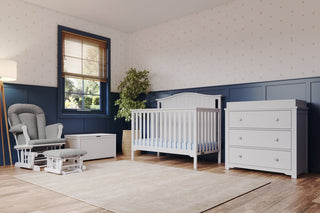A cozy nursery room with a white crib, matching dresser, storage bench, and the Child Craft Cozy Glider and Ottoman in matte white. Features large window, potted plant, blue wainscoting, and patterned wallpaper.