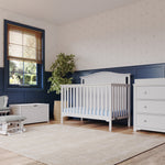 A cozy nursery showcases the Child Craft Hampton Arch Top 4-in-1 Convertible Crib in matte white, paired with a matching dresser and storage bench. A gray cushioned rocker, blue wainscoting, beige rug, and a potted plant complete the space.