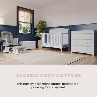 A cozy nursery features a matte white Cozy Glider and Ottoman by Child Craft, white crib, matching dresser, beige rug, blue and white beadboard walls with light wallpaper, and large windows. Text: “Classic Cozy Cottage.”.