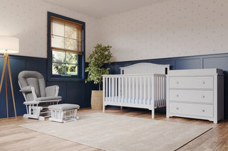 A cozy nursery with a Child Craft Cozy Glider and Ottoman in Matte White, a white crib, matching dresser, blue wainscoting, patterned wallpaper, large window, floor lamp, potted plant, and light rug.