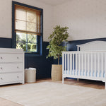 A bright nursery features the Child Craft Hampton Arch Top 4-in-1 Convertible Crib in Matte White with adjustable mattress support, a matching dresser, potted plant, wooden blinds, blue and white walls, and a beige rug on wood floors.
