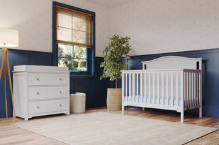 A bright nursery features the Child Craft Hampton Arch Top 4-in-1 Convertible Crib in Matte White with adjustable mattress support, a matching dresser, potted plant, wooden blinds, blue and white walls, and a beige rug on wood floors.
