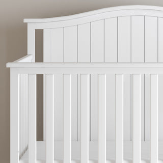 A close-up of the Child Craft Hampton Arch Top 4-in-1 Convertible Crib in Matte White, with vertical slats, a curved headboard, and adjustable mattress support, set against a neutral wall.