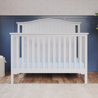 The Child Craft Hampton Arch Top 4-in-1 Convertible Crib in matte white stands on a wooden floor, against blue wainscoting and light wallpaper. It features adjustable mattress support and a blue star-patterned mattress, with a basket and plant nearby.