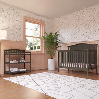 A bright nursery features the Child Craft Hampton Arch Top 4-in-1 Convertible Crib in Dapper Grey, a matching changing table with baby items and plush toy, large potted plant, geometric rug, pink paneled walls, dotted wallpaper, and a sunlit window.