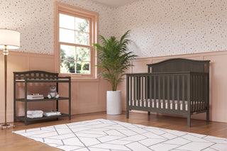 A bright nursery features the Child Craft Hampton Arch Top 4-in-1 Convertible Crib in Dapper Grey with adjustable mattress support, a dark wood changing table, white geometric rug, potted plant, lamp, window, polka dot pink walls, and a stuffed animal on the shelf.