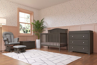 A modern nursery features gray furniture, including the Child Craft Cozy Glider and Ottoman in Dapper Gray, a crib, dresser, and toy chest. The room includes pink paneling, dotted wallpaper, a large window, plant, and geometric rug.