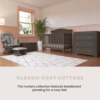 A nursery with gray furniture features the Child Craft Hampton Arch Top 4-in-1 Convertible Crib in Dapper Grey, a dresser, rocking chair, white rug, potted plant, window with pink trim, beadboard paneling, and dotted wallpaper.