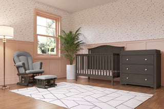 A modern nursery with the Child Craft Hampton Arch Top 4-in-1 Convertible Crib in Dapper Grey, adjustable mattress support, matching dresser, gray glider with ottoman, geometric rug, tall plant, large window, and pink dotted wallpapered walls.
