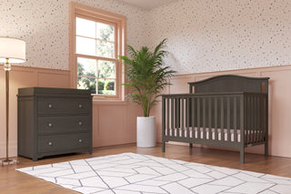 A nursery with a Child Craft Hampton Arch Top 4-in-1 Convertible Crib in Dapper Grey, matching dresser, white potted plant, floor lamp, geometric rug, wood flooring, pink paneled walls, speckled wallpaper, and sunlight through the window.