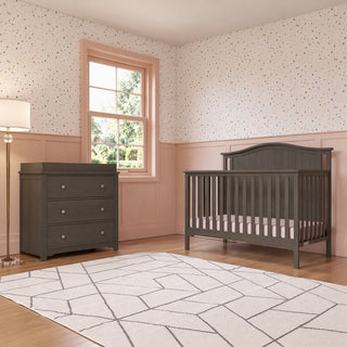 A dark gray Child Craft Hampton Arch Top 4-in-1 Convertible Crib and a matching three-drawer dresser in a nursery with pink wainscoting and terrazzo-patterned wallpaper.